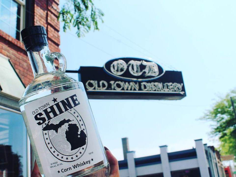 Bottle of OTD's Old Town Shine held up in front of the Old Town Distillery sign in Old Town Saginaw
