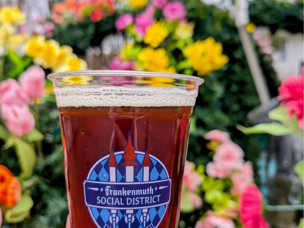 Person holding a to-go beer from Frankenmuth Brewery in a Frankenmuth Social District cup in front of colorful flowers in the outdoor Frankenmuth Social District in Frankenmuth, Michigan