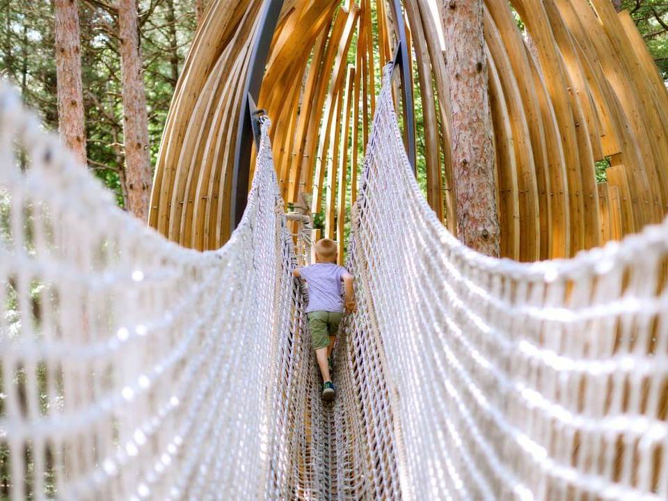Boy crossing the cargo net bridge to a wooden pod play structure along the Whiting Forest of Dow Gardens Canopy Walk