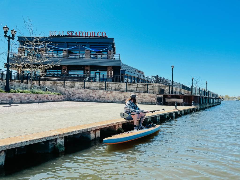 Guy sitting along the Saginaw River in Bay City with his stand-up paddleboard by Real Seafood Company in Uptown