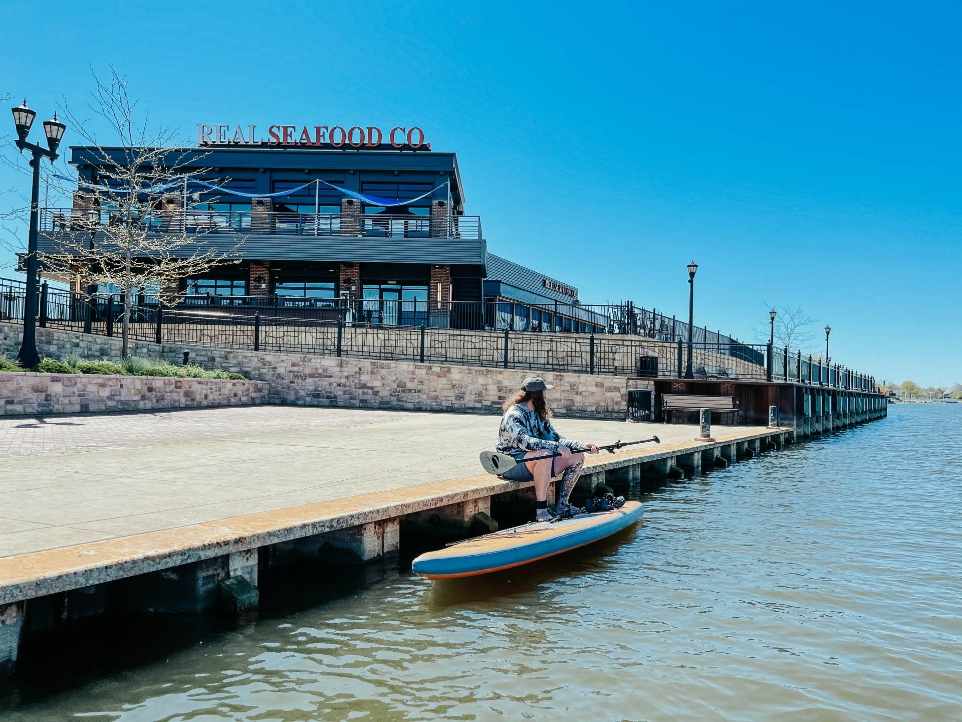 Guy sitting along the Saginaw River in Bay City with his stand-up paddleboard by Real Seafood Company in Uptown